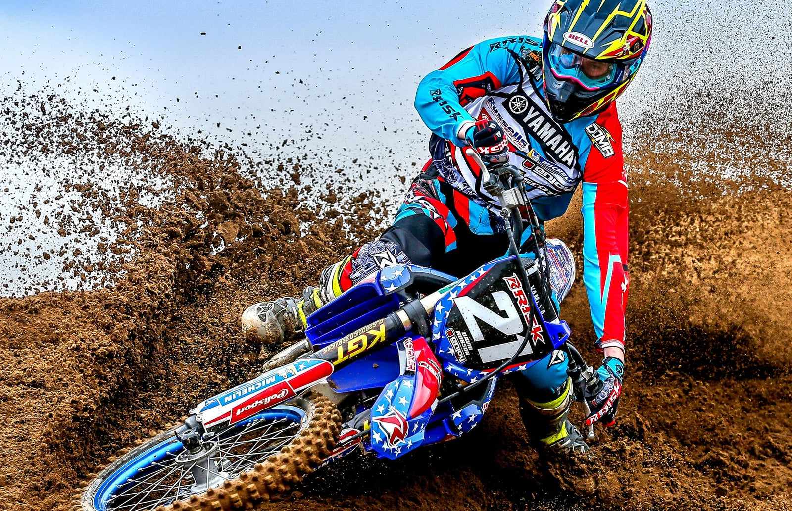 Motocross racer hitting a berm with loose dirt while on the throttle causing dirt to spray behind him as they ride through the turn? The rider has risk racing grips installed on their bike and are wearing risk racing motocross gear.